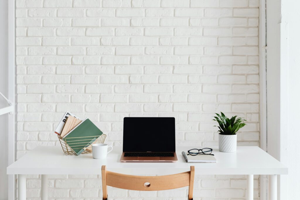 Clean and organized white workspace with laptop, plant, and books against a brick wall.